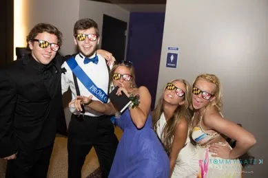 Five people wearing formal attire and colorful sunglasses pose playfully for the camera in a hallway.