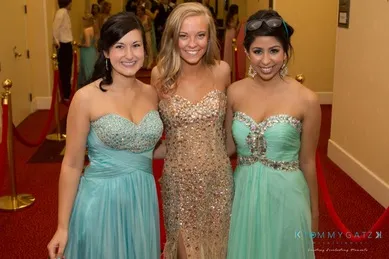 Three women in evening gowns pose together at an event, standing on red carpet flooring. Two wear aqua dresses, and the woman in the center wears a shimmering gold dress.