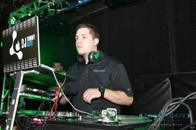 DJ performing at a set with headphones around his neck, standing behind a laptop and mixing equipment on stage with green lighting.