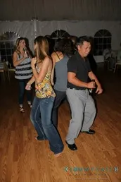 People dancing energetically on a wooden floor. One person holds a drink. Indoor setting with others in the background.