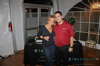 Two people standing together in front of a DJ booth with a Tommy Gatz Events banner. The woman is holding a drink, and both are smiling at the camera.