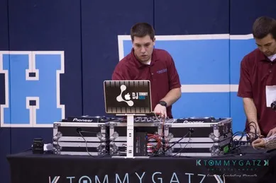 DJ operating equipment at an event, standing next to another person. Both are wearing maroon shirts. Background features blue wall with large letter "H".