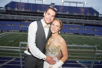 A couple dressed formally stand together at a stadium, smiling at the camera. The field is empty in the background.