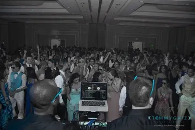 A large crowd dances in a dimly lit event hall, with a DJ booth in the foreground.