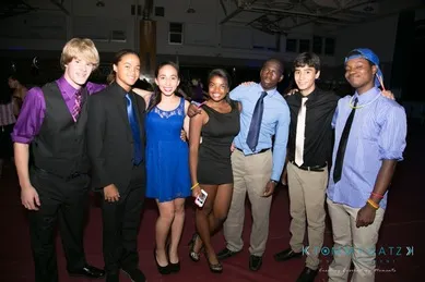 A group of seven young people dressed in semi-formal attire are posing together indoors at an event.
