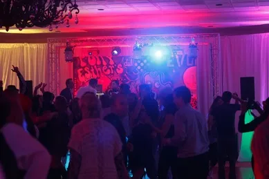 A crowded dance floor with people dancing under colorful lights. A graffiti-style backdrop is visible in the background.