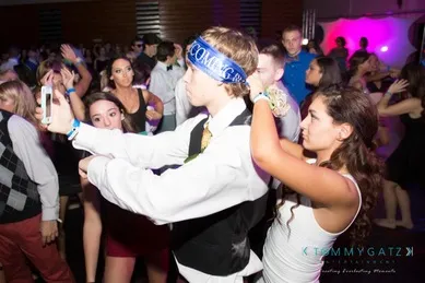 Teens dancing at a prom, one wearing a "Homecoming" sash, taking a selfie. Crowded room with colorful lights in the background.