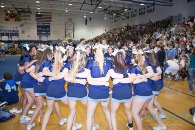 A group of cheerleaders in blue uniforms huddle on a gym floor during a school event, surrounded by spectators on bleachers.