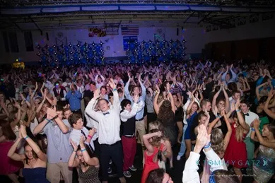 A large group of people, dressed in formal attire, dance energetically at a crowded indoor event with blue star decorations on the wall.