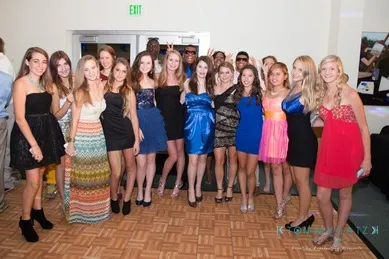 A group of people posing together, most wearing dresses and standing on a parquet floor in a brightly lit room.