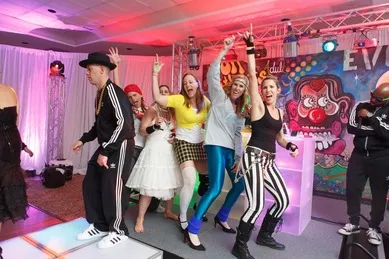 A group of people in colorful costumes dance and pose enthusiastically on a stage with vibrant lighting and a painted backdrop.