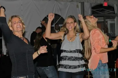 Three women are dancing and smiling energetically at a party in a room with a white ceiling and drapes.