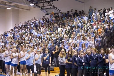 A large crowd of people, including students in sports uniforms, fill bleachers in a gymnasium setting.