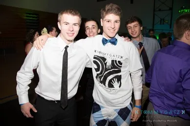 A group of four young men posing together at an indoor event. One wears a graphic T-shirt and bow tie, while the others are in formal attire with ties.