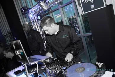 A DJ wearing a black shirt is operating equipment at a music event. There's a laptop and mixer on the table, and blue lighting in the background.