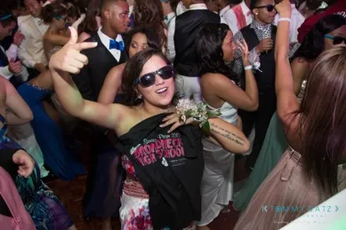 A group of people dance at a party. The woman in the foreground wears sunglasses and holds a shirt, smiling and gesturing with her hand.