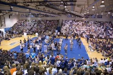 A large crowd in a gym watches a band perform at the center of the blue floor, surrounded by seated spectators.