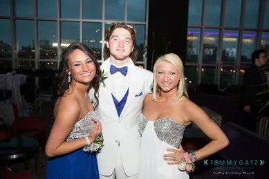 Three people dressed in formal attire pose together inside a room with large windows.