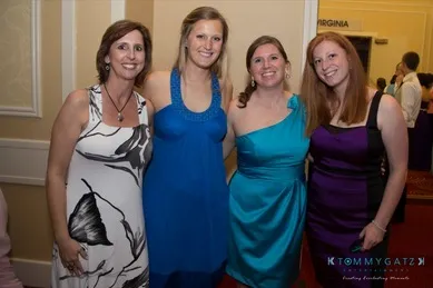 Four women smiling and posing together in a hallway, each wearing a different colored dress.