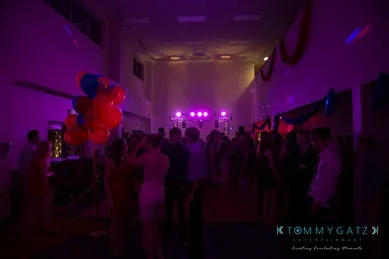A dimly lit party with people dancing. Bright pink and purple lights illuminate the room, which is decorated with red and blue balloons.