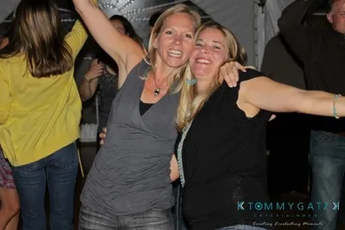 Two women smiling and posing with arms around each other at a lively indoor gathering, with other people in the background.
