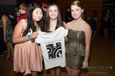 Three young women pose at an event; one holds up a T-shirt with a graphic design.