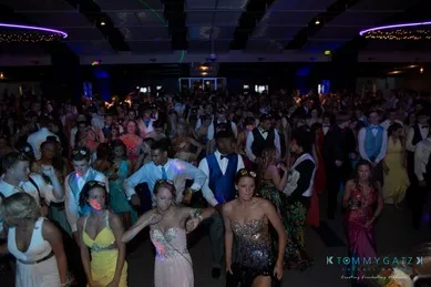 A large group of people in formal attire dance in a dimly lit event space with colorful lights overhead.