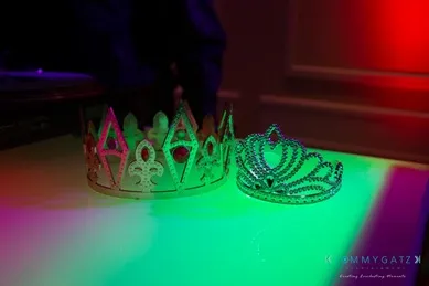 Two crowns rest on a surface under colorful lighting; one is gold with triangular peaks, the other is silver with a fan-like design.