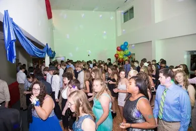 A large group of people in formal attire stand in a brightly lit room with colorful balloons in the background.