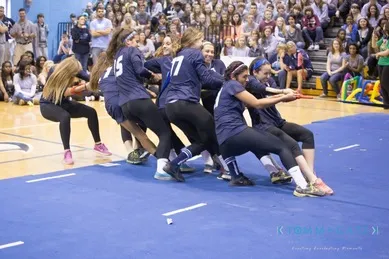 A team of women in matching sports attire participates in a tug-of-war competition inside a gym with a crowd watching.