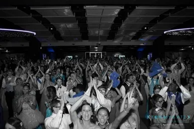 A large group of people in formal attire celebrate and clap at an indoor event, with dim lighting and a colorful highlight on their outfits.