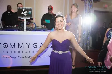 A woman in a purple dress and tiara smiles with her arms open. A DJ and three other people are in the background near a sound system.