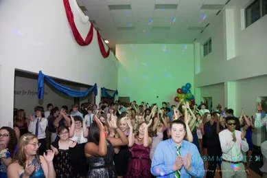 A large group of people in formal attire are clapping and dancing at an indoor event decorated with blue and red drapes and balloons.