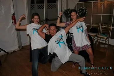 Three people posing energetically with flexed arms. Two women wear white T-shirts over their clothes, while a man crouches in front, pointing with one arm.