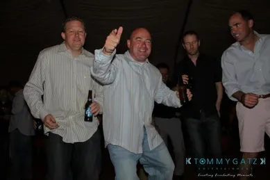 Five men are standing together, holding bottles, with one man smiling and pointing toward the camera.