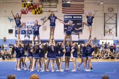 Cheerleading team in blue uniforms performs a pyramid stunt in a gymnasium with banners and flags in the background.