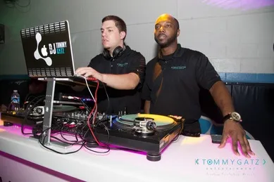 Two DJs standing behind a DJ booth with equipment, one wearing headphones and using a laptop while the other looks on, in a dimly lit setting.