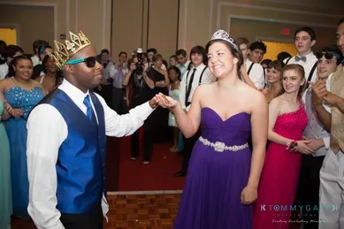 A couple in formal attire, both wearing crowns, dance at a prom event. The woman is in a purple gown, and the man wears a blue vest and sunglasses. People in formal wear are in the background.