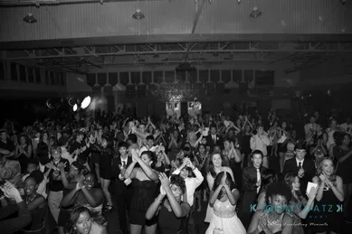 A large group of people dressed formally, clapping and cheering in a dimly lit venue with chandeliers and a high ceiling.