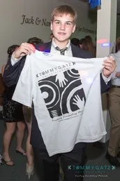 A young man in a suit holds up a white T-shirt with a graphic design on it.