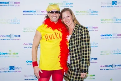 A person in a yellow outfit with "HULKA" stands next to a person in a plaid suit. They are smiling at an event with a branded backdrop.