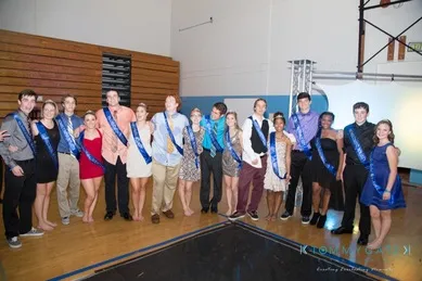 A group of people wearing blue sashes stand together in a gymnasium, posing for a photo. Some are dressed formally while others are in casual attire.