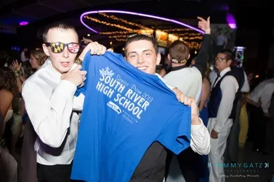 Two young men at a dance, one holding a "South River High School" shirt. The venue has dim lighting with decorative lights in the background.