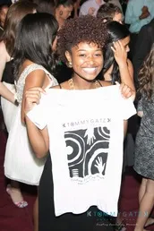 A person smiling and holding a white T-shirt with a graphic design at an indoor event.