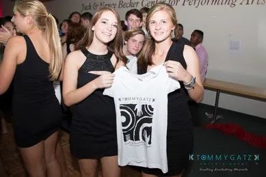 Two women in black dresses hold a white T-shirt at an indoor gathering. People in the background appear engaged in conversation.
