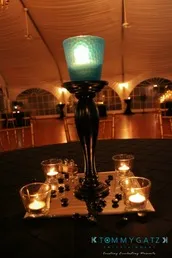 Tall black candle holder with a blue candle, surrounded by small tea lights set on a dark tablecloth. Background shows a draped ceiling and dimly lit room.
