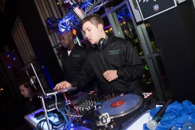 A DJ wearing headphones is spinning records at a music event. Another person is in the background near the equipment.