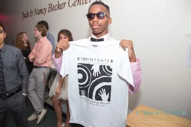 A person wearing sunglasses holds a white T-shirt with abstract graphics and text. Other people are in the background near a wall with text.