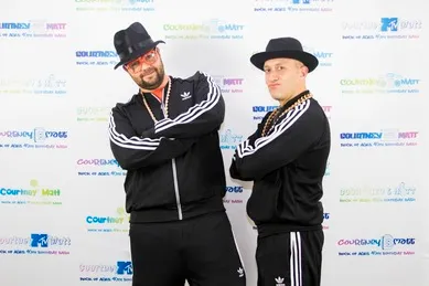 Two people in matching black track suits and hats stand with arms crossed against a branded backdrop.