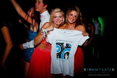 Two smiling women at a party holding a white t-shirt with a blue and black design. People in formal attire are dancing in the background.